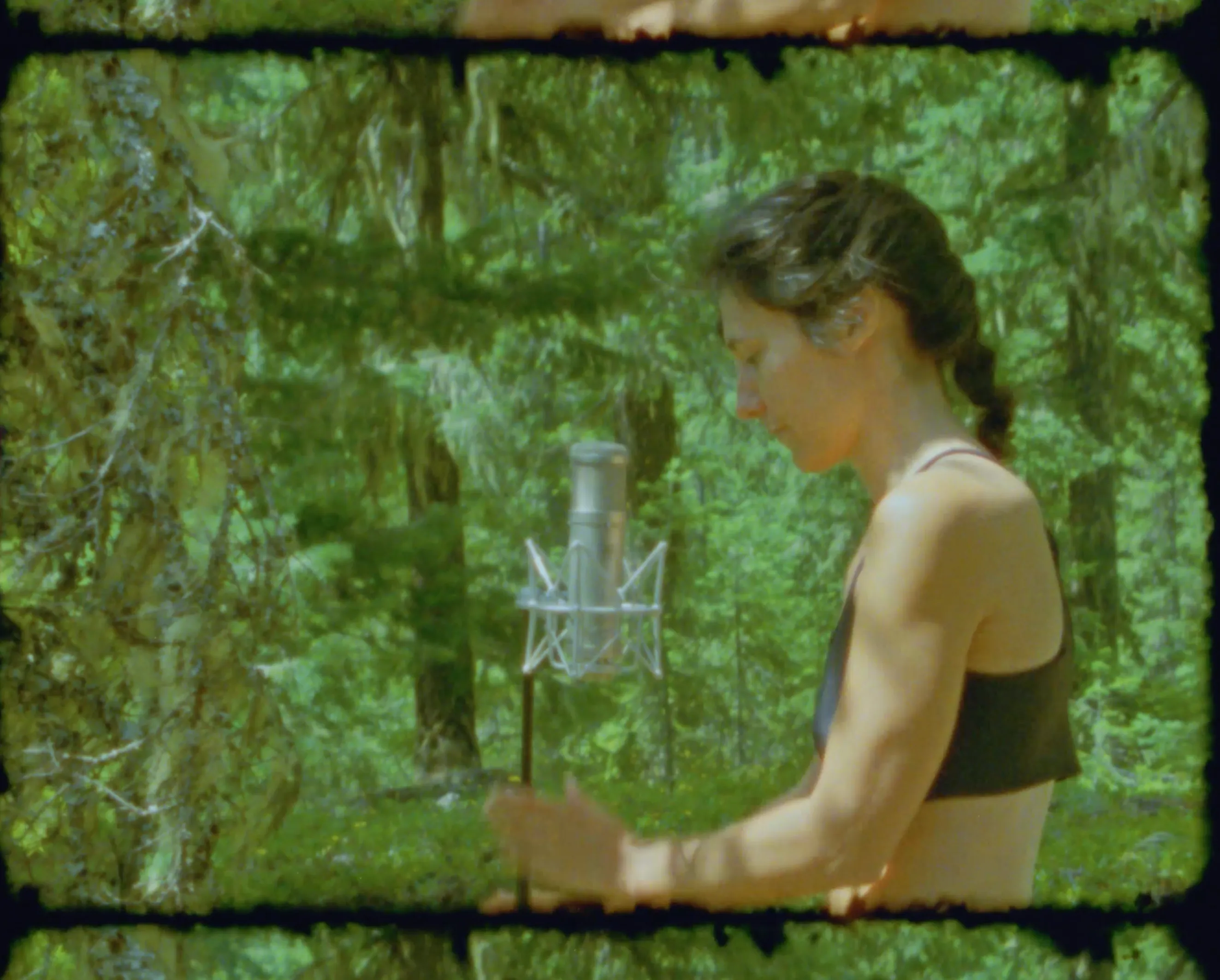 A vocalist stands at a large condenser microphone in a shock mount, surrounded by deep green forest